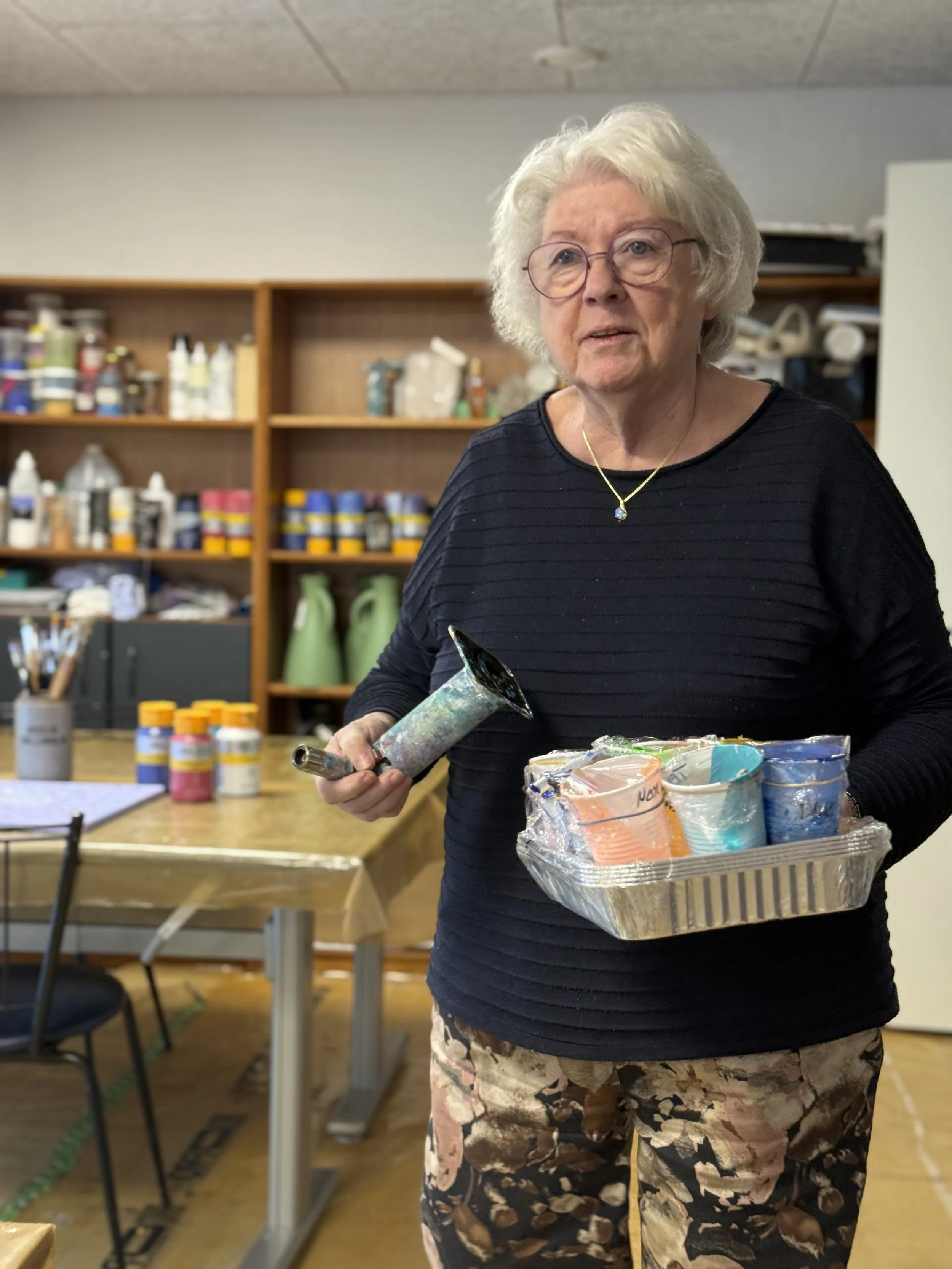 En ældre kvinde med hvidt, krøllet hår og briller står i et værksted og holder en plastbakke med farvede kopper og et luftværn. Hun er iført en sort langærmet top og bukser med blomsterprint.