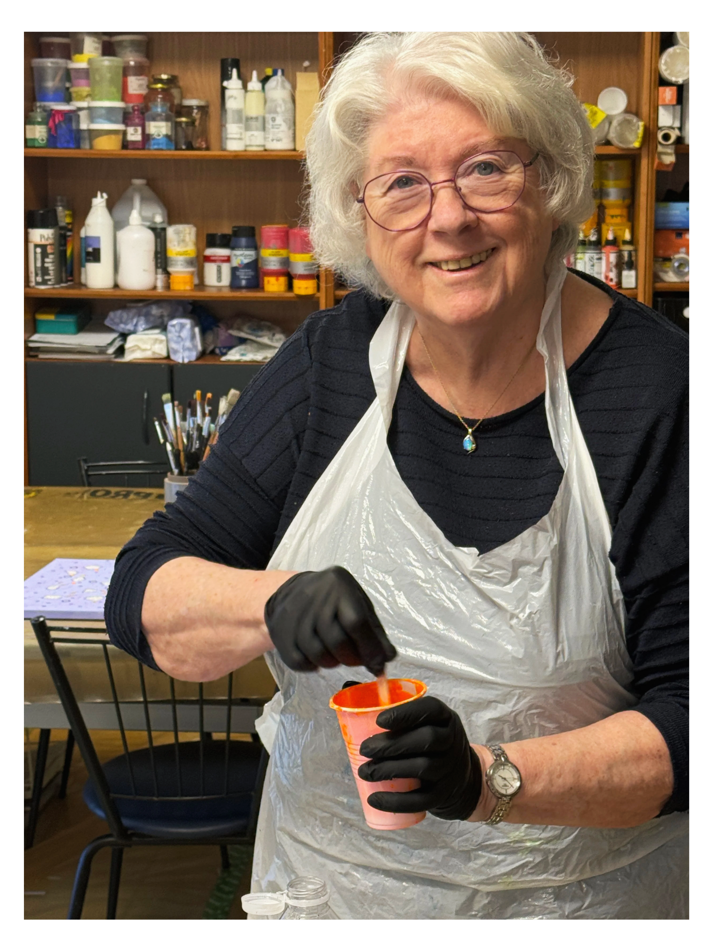 En ældre kvinde med brunt, intet hår og briller, der smiler og hælder farvet maling i en kop. Hun bærer handsker, en hvid plasticforklæde, en sort langærmet trøje, et ur og et smykke med en blå sten. Bag hende er der hylder med forskellige lakker og materialer.