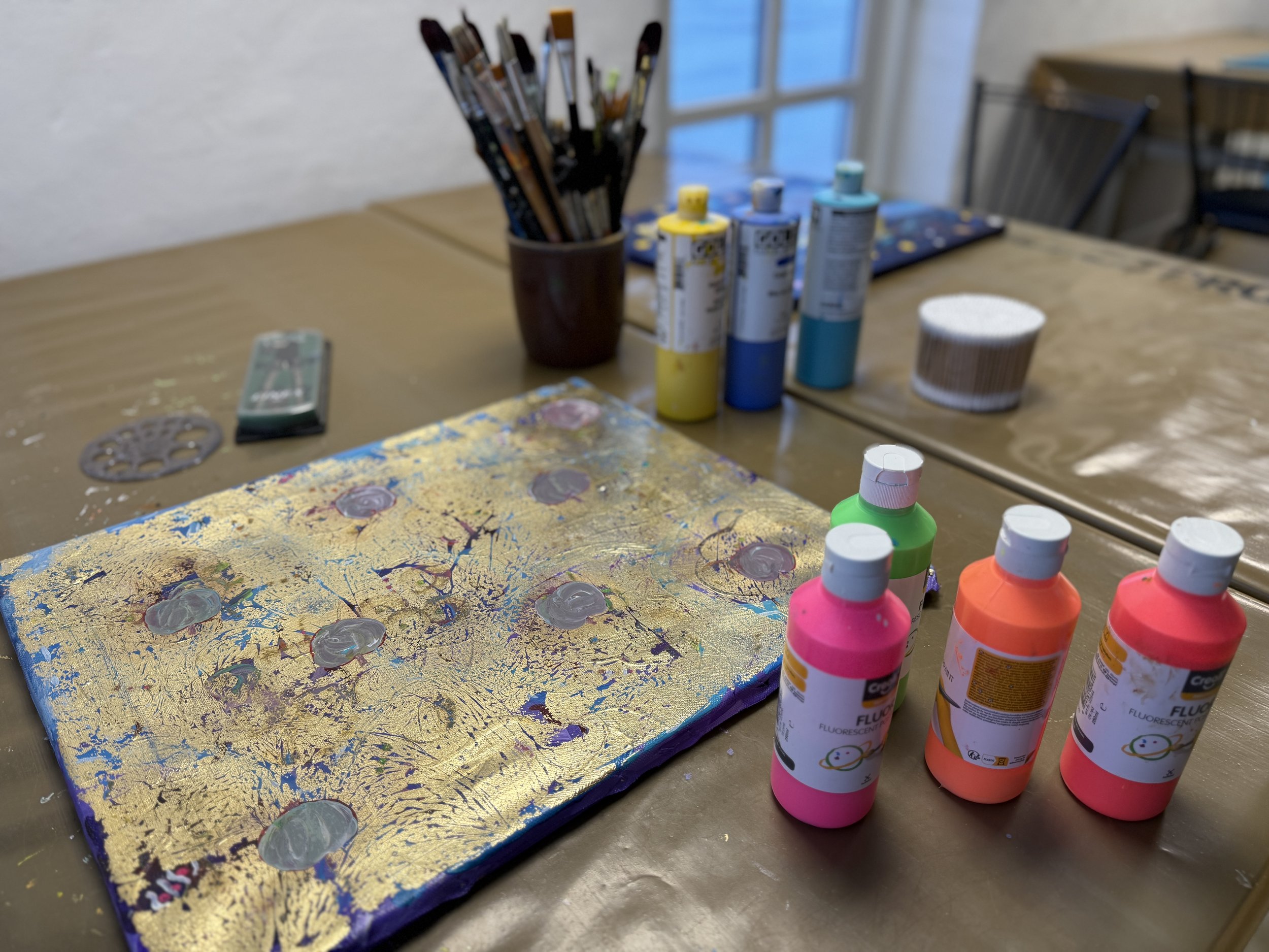 Et bord med kunstnermaterialer, herunder maling, flasker med fluorescerende maling, pensler i en kop, og en malet lærred med guld, lilla og blå akrylfarver.