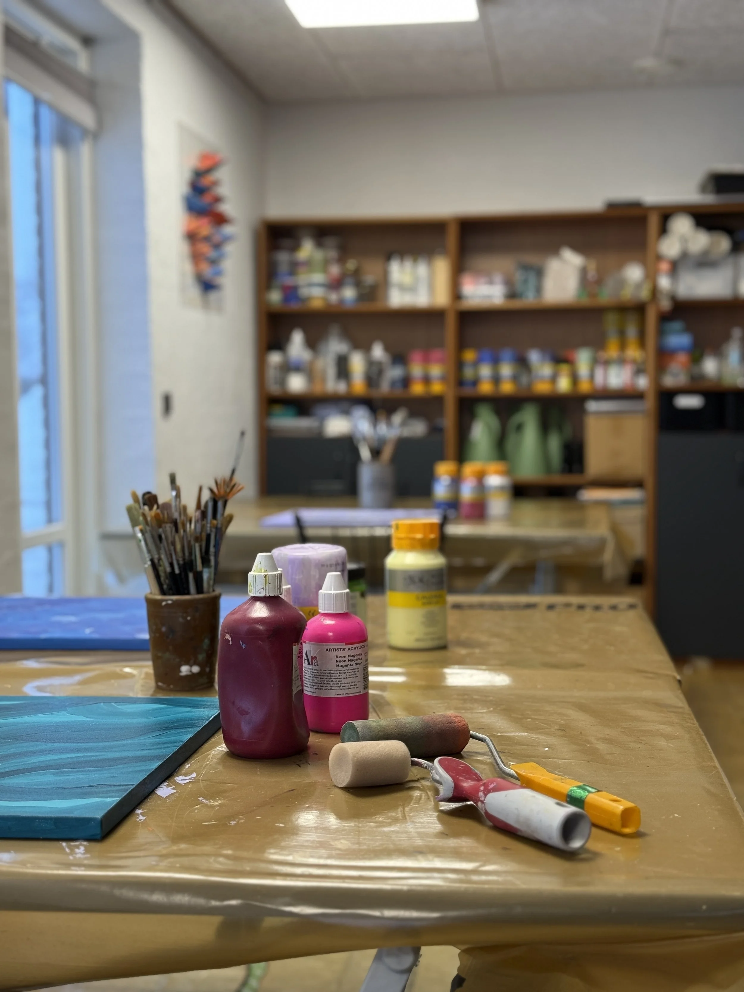 Et kunstnerisk værksted med maling og maleudstyr på et bord, med opbevaringsreol fyldt med materialer i baggrunden.