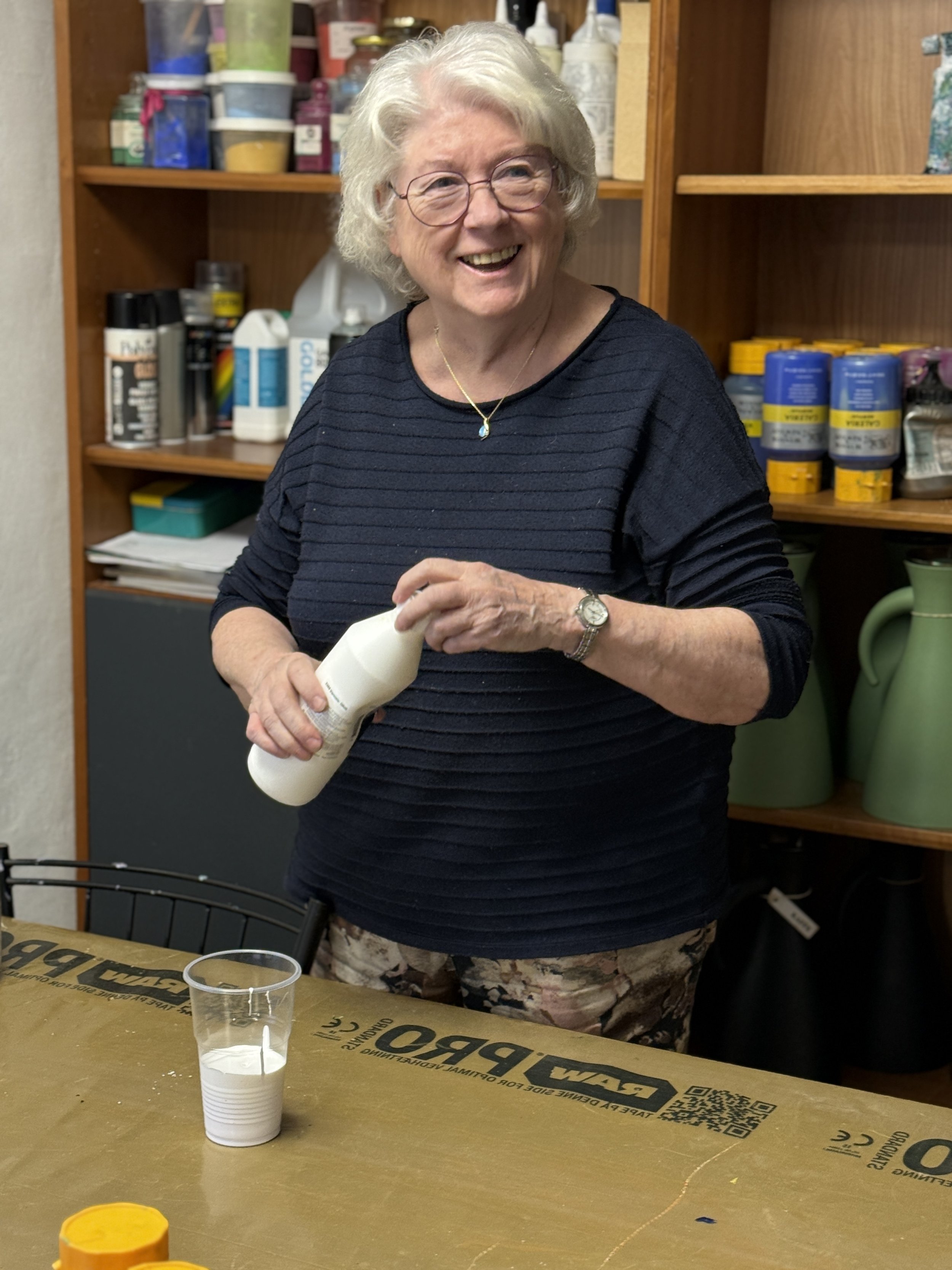 En ældre kvinde med lyst, krøllet hår og briller smiler, mens hun holder en hvid plastflaske. Hun står bag et bord med en glas med et hvidt pulver og andre genstande, i et rum med hylder fyldt med diverse kunstnermaterialer og forsyninger.
