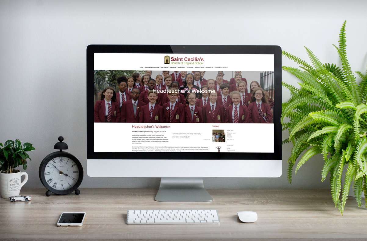 Computer monitor displaying the homepage of Saint Cecilia's Church of England School website, with a group photo of students in school uniforms and the headline "Headteacher's Welcome." The workspace includes a clock, potted plants, a smartphone, a k