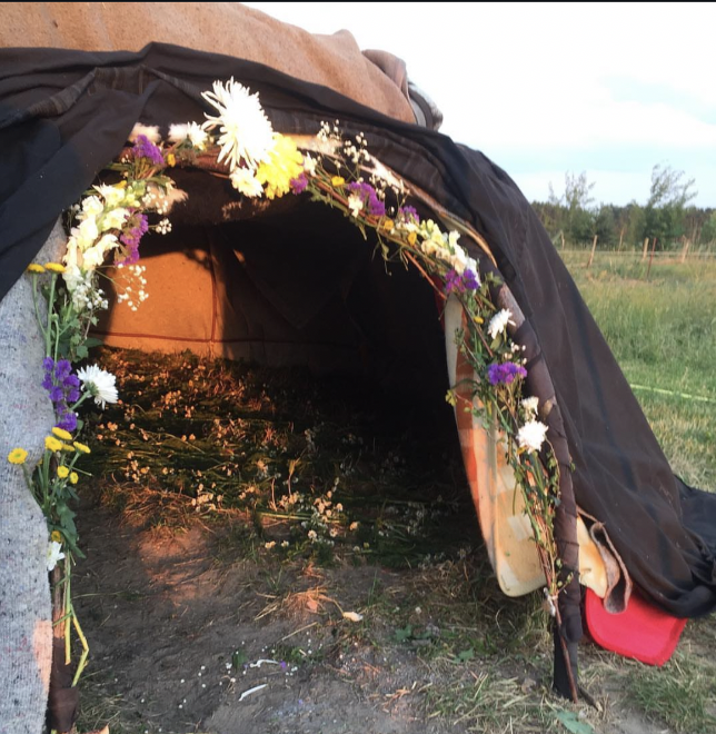 The entrance to a dark ceremonial sweat lodge, draped in purple and yellow wildflowers