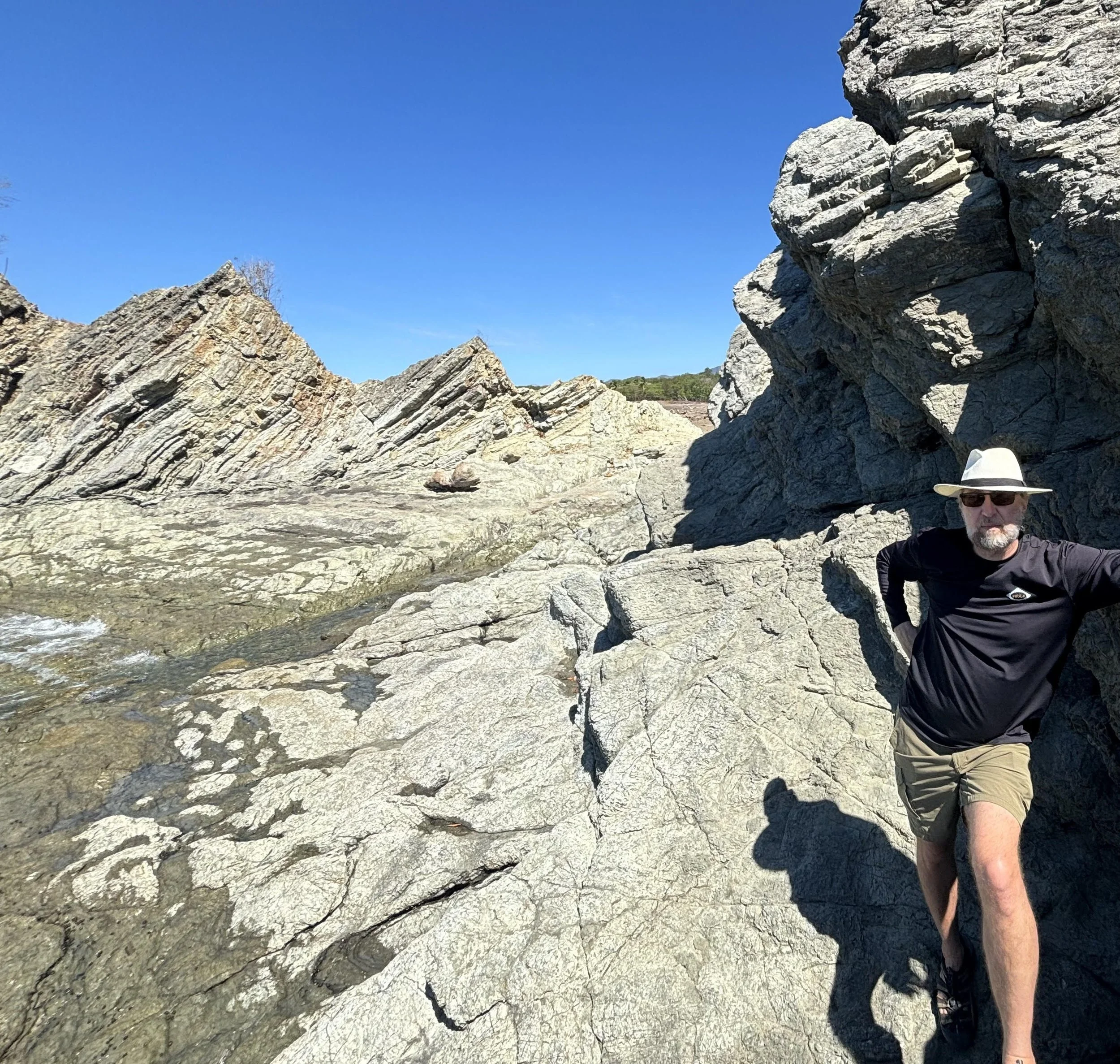 This picture shows a middle-aged white man with dark glasses, a white hat, and a beard standing amid rocky Costa Rican cliffs.