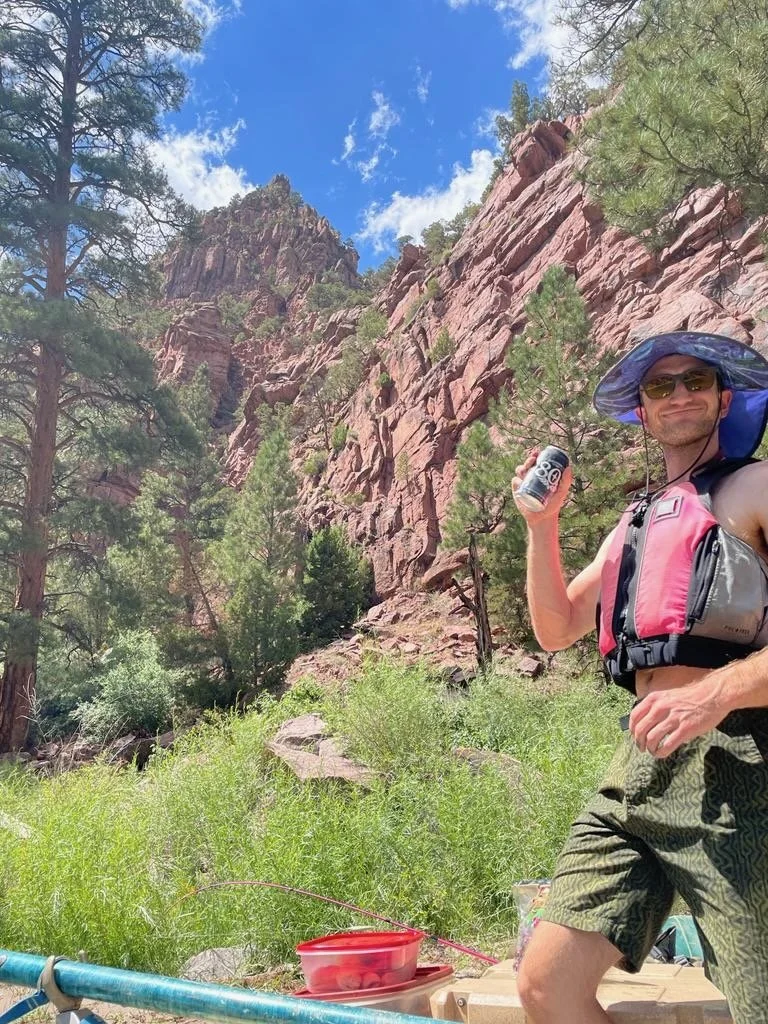 The writer Derek Maiolo stands under pine trees and a wall of sandstone while rafting the Green River in Utah