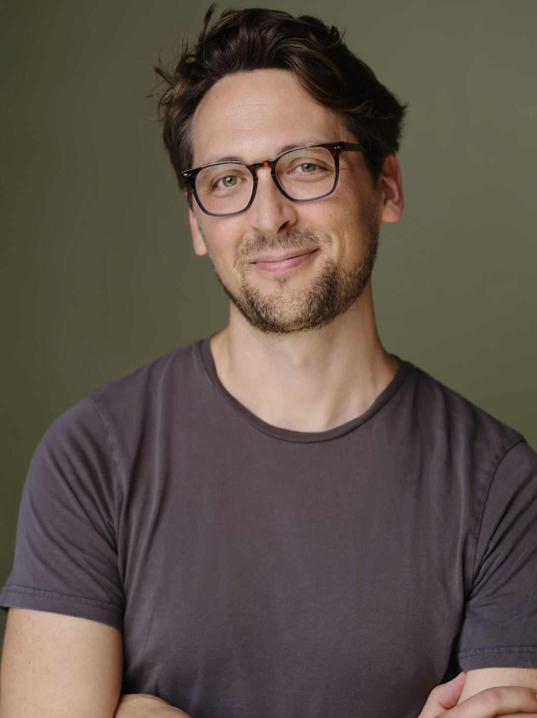 A slightly unshaven man with short wavy hair, a gray t-shirt, and glasses half-smiles at the viewer.