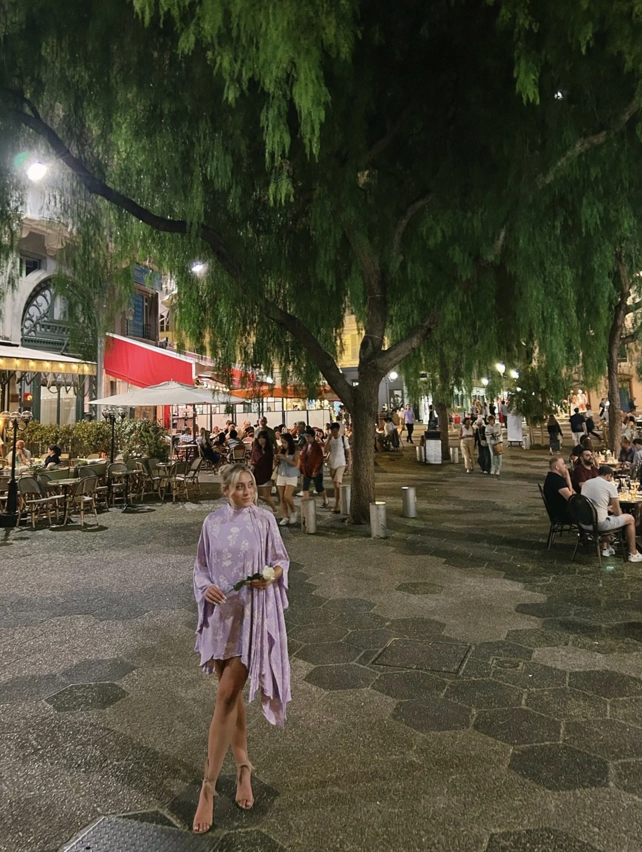 A blonde woman wearing a purple dress stands in a French courtyard holding a white rose and peering off to the side.