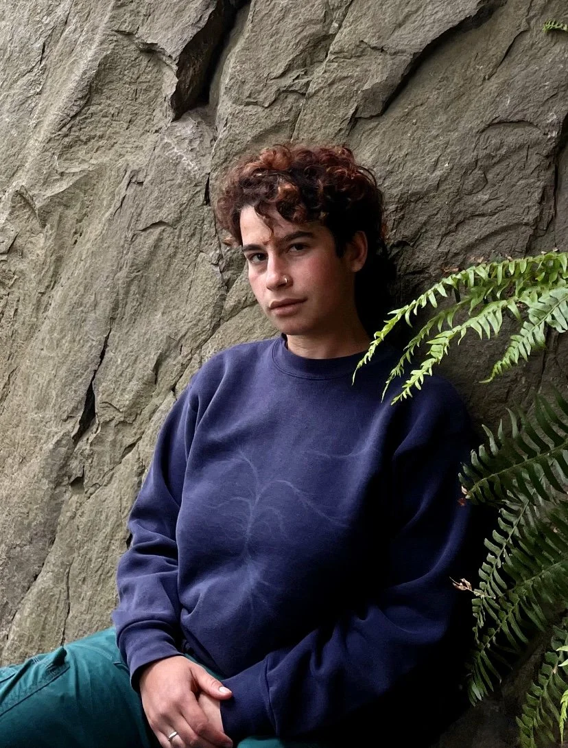 A photo of the author, wearing a dark blue sweatshirt, sitting outside next to a rock wall and a fern plant.