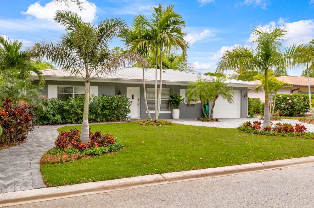 A single-story house with a gray exterior, white front door, and garage, surrounded by lush green lawns, palm trees, and tropical plants on a bright, sunny day.