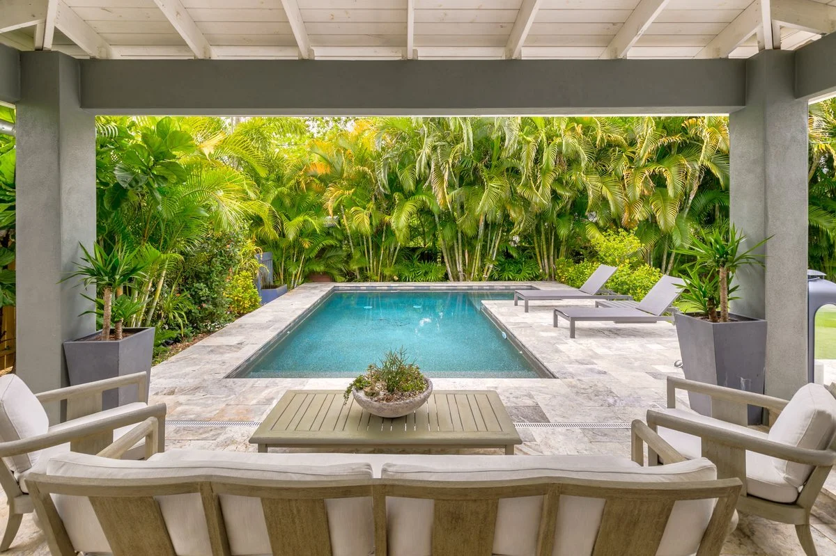 Private backyard with a swimming pool surrounded by tropical plants and lounge chairs, viewed from a covered patio area with outdoor furniture.