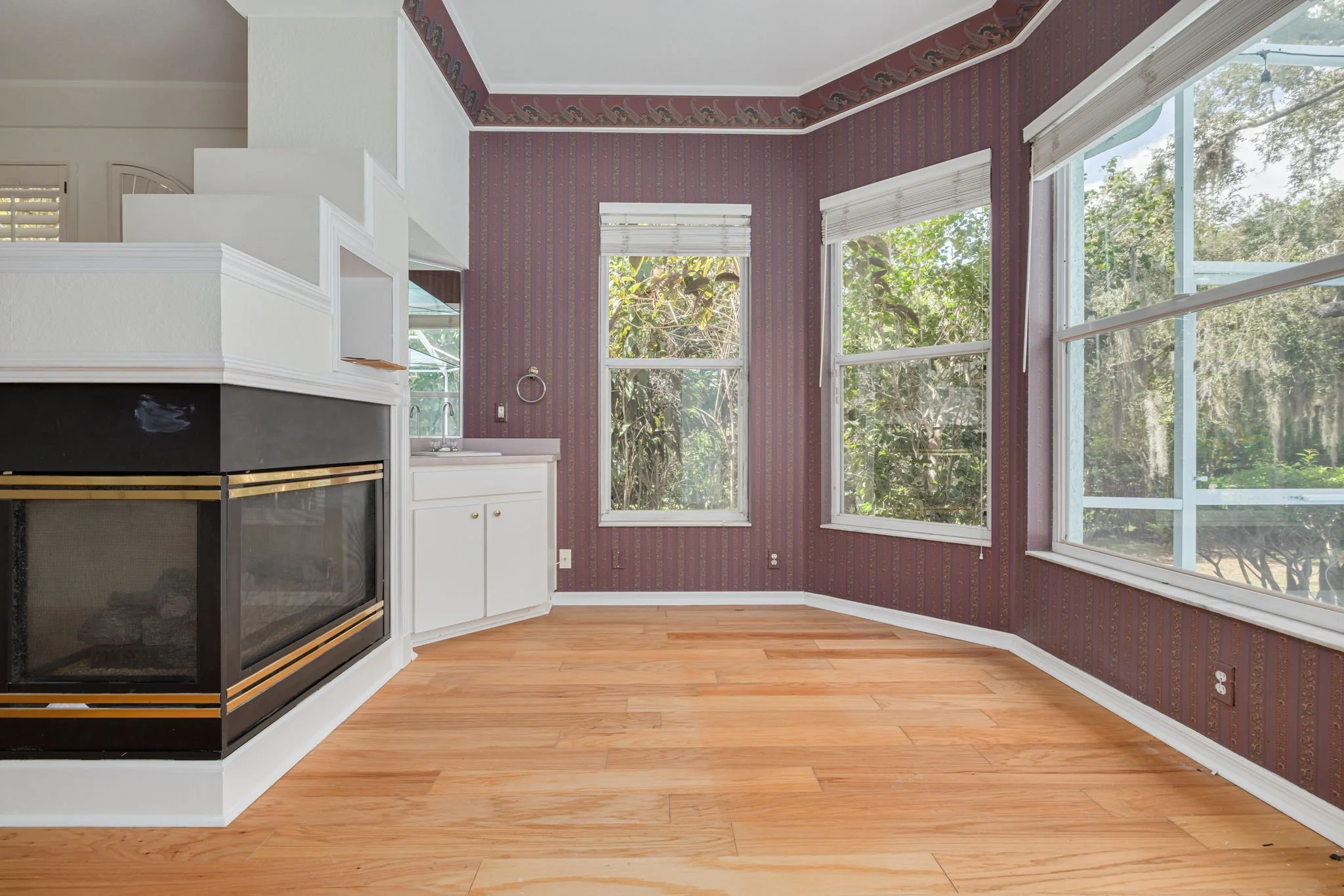 A corner of a room with purple patterned wallpaper, hardwood floor, and large windows showing trees outside. There is a fireplace with a black and gold trim, white cabinetry, and a small sink area nearby.