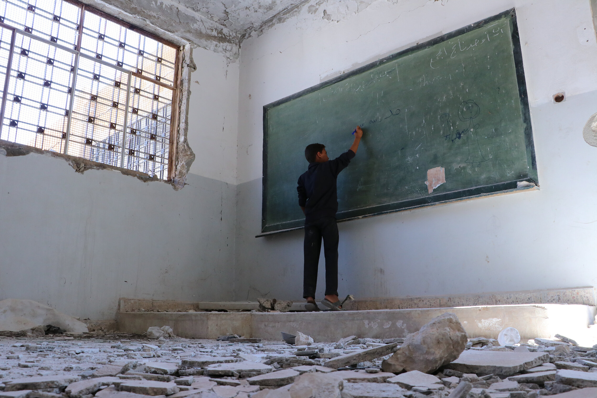 A boy writes on the board of a damaged classroom in Idlib governorate, Syria in July 2019. © 2019 Save the Children's partner in Syria, Hurras Network.