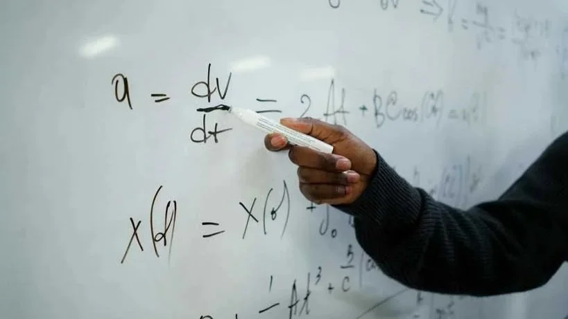 Photo of a whiteboard covered with mathematical equations. We can see a black hand, wearing a dark jumper, underlining one of the equations with a pen.