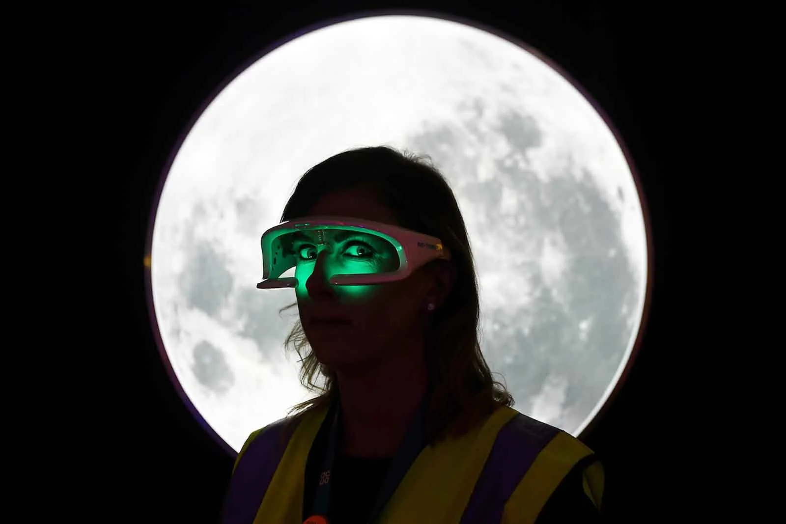A woman wearing 3D glasses with green light, standing in front of the moon, silhouette visible.