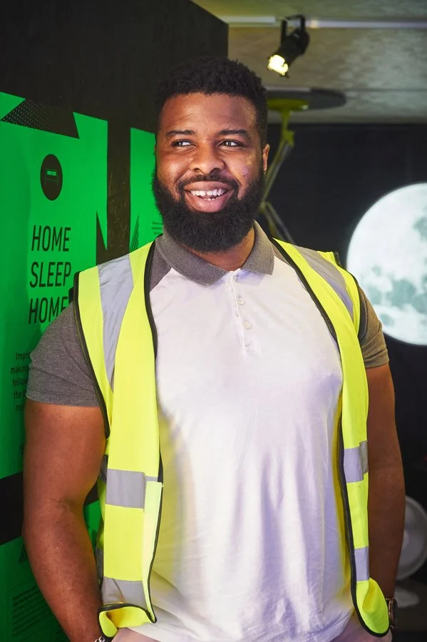 Dr Ron Dodzro. A smiling man with a beard and short curly hair wearing a grey polo shirt and a bright yellow safety vest standing indoors, behind him are green signs and a large image of the moon.