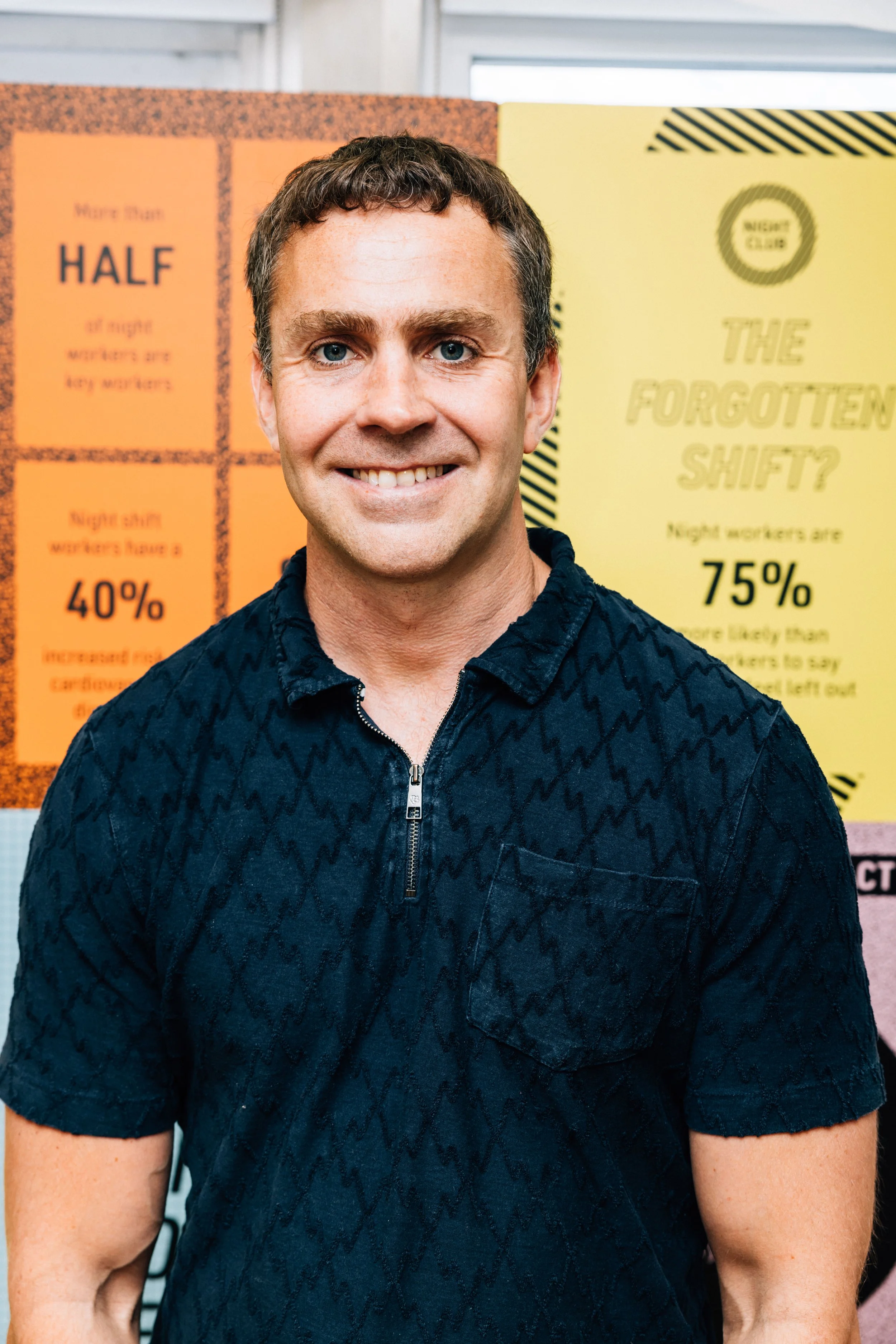 A photo of Dr Steve Walsh. A smiling man with short brown hair and blue eyes wearing a dark patterned zip-up shirt, standing in front of informational posters about night shift workers.