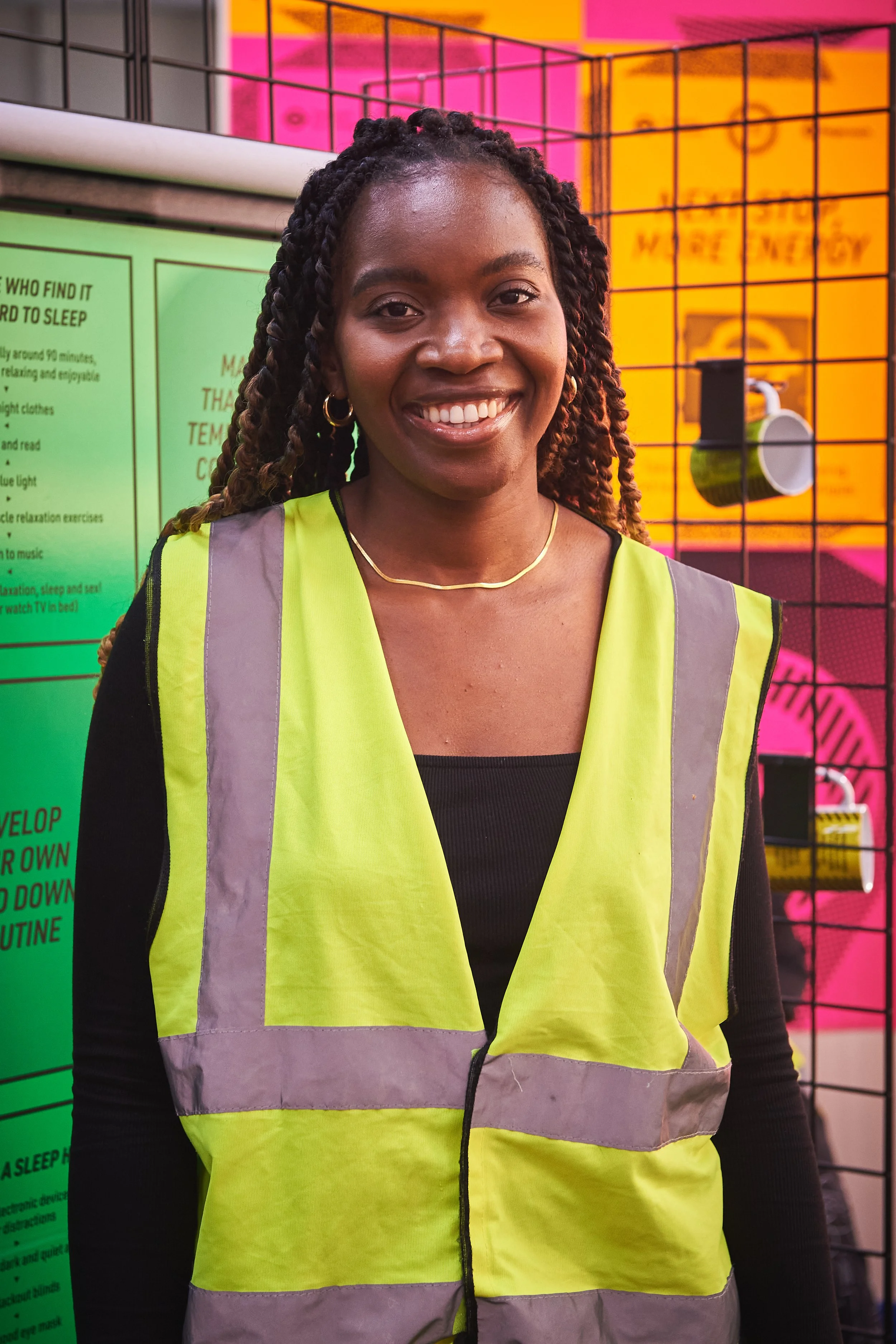 A photo of Dr Sarah Atayero. A smiling woman wearing a yellow safety vest with reflective strips, standing in front of colourful display panels of the Night Club Flex. 