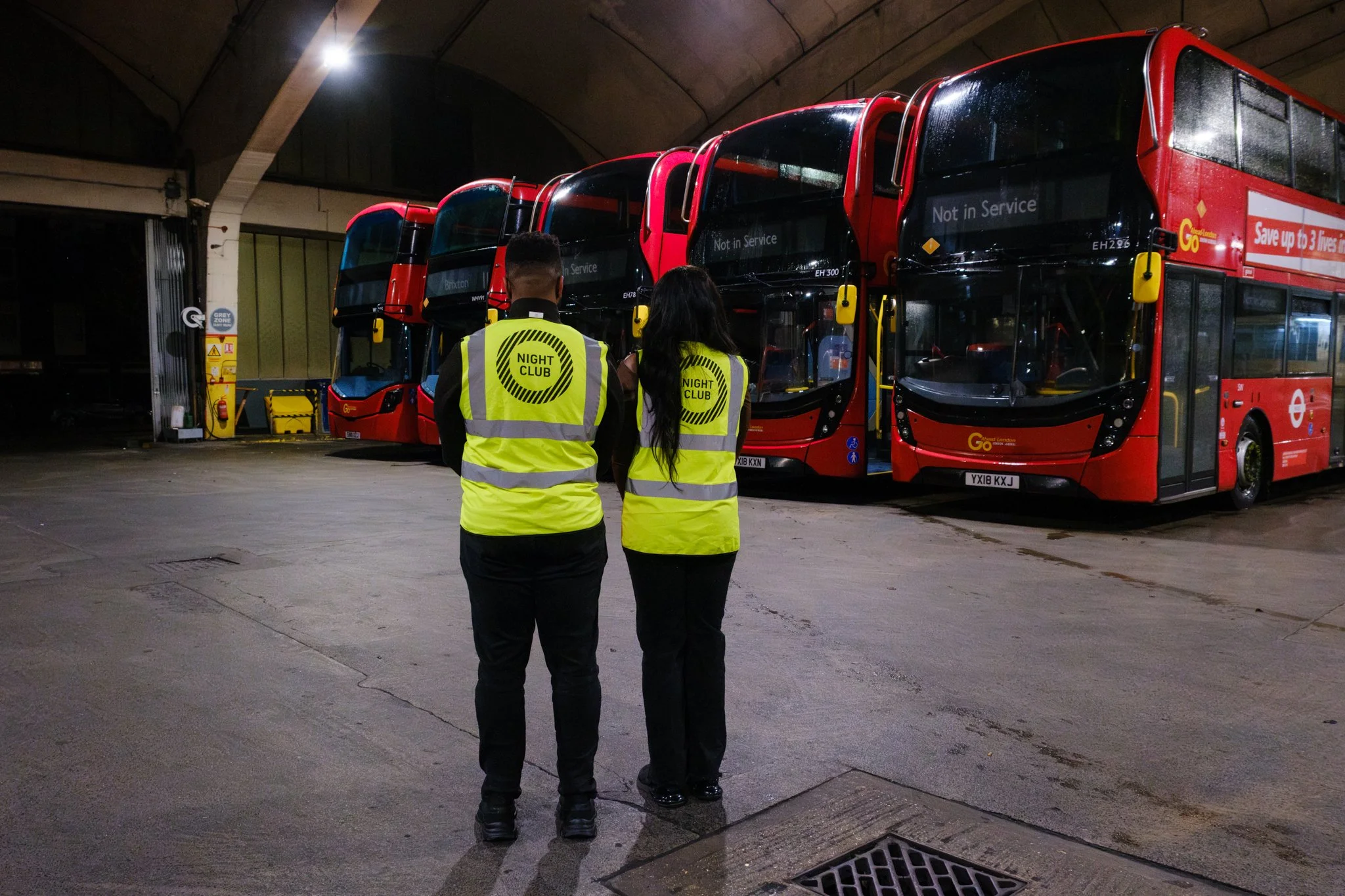 Two people in high vis vests showing the Night Club logo on their backs stand in front of Transport for London buses in a depot