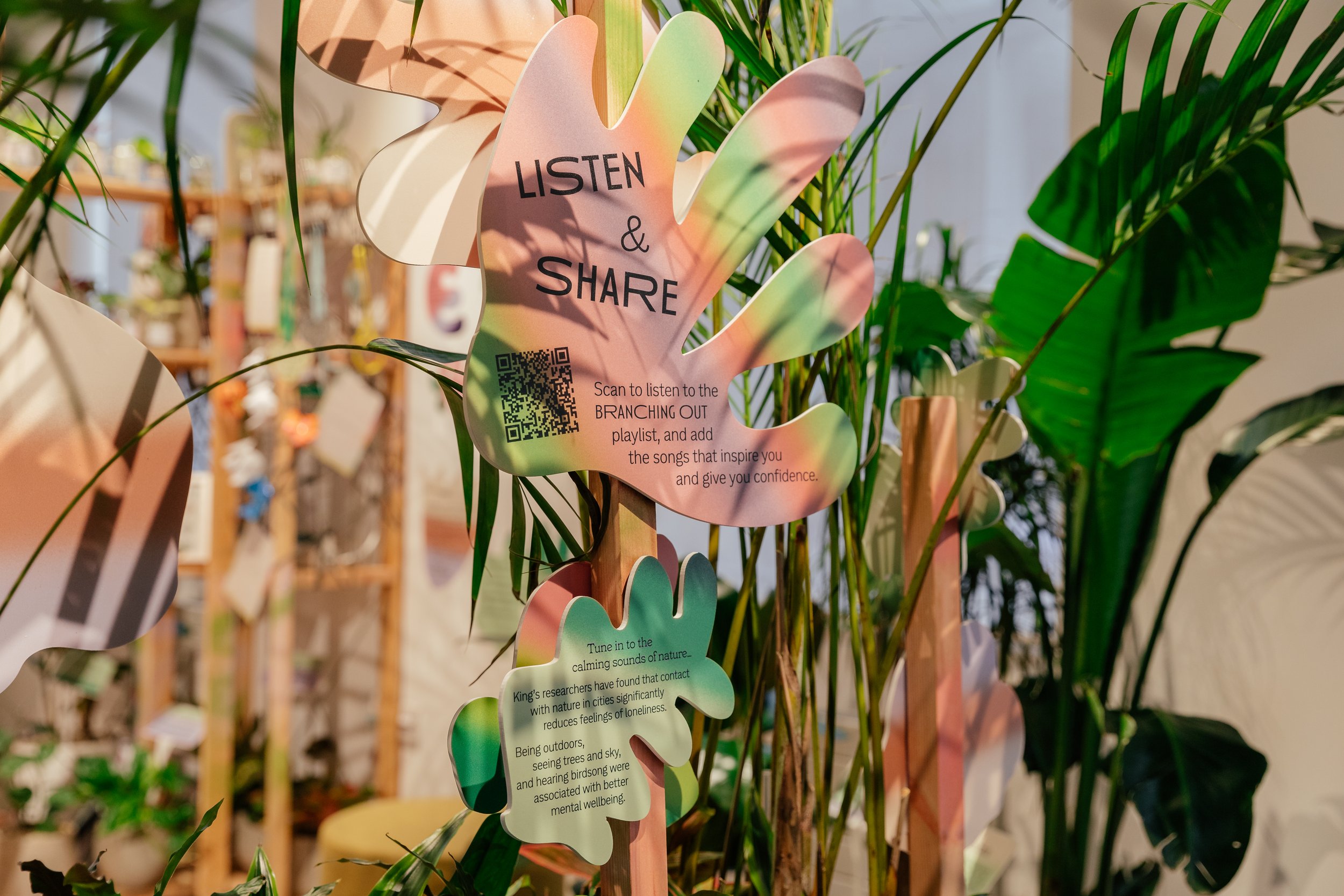 Colorful leaf-shaped sign with the words 'Listen & Share' and a QR code, surrounded by green plants.