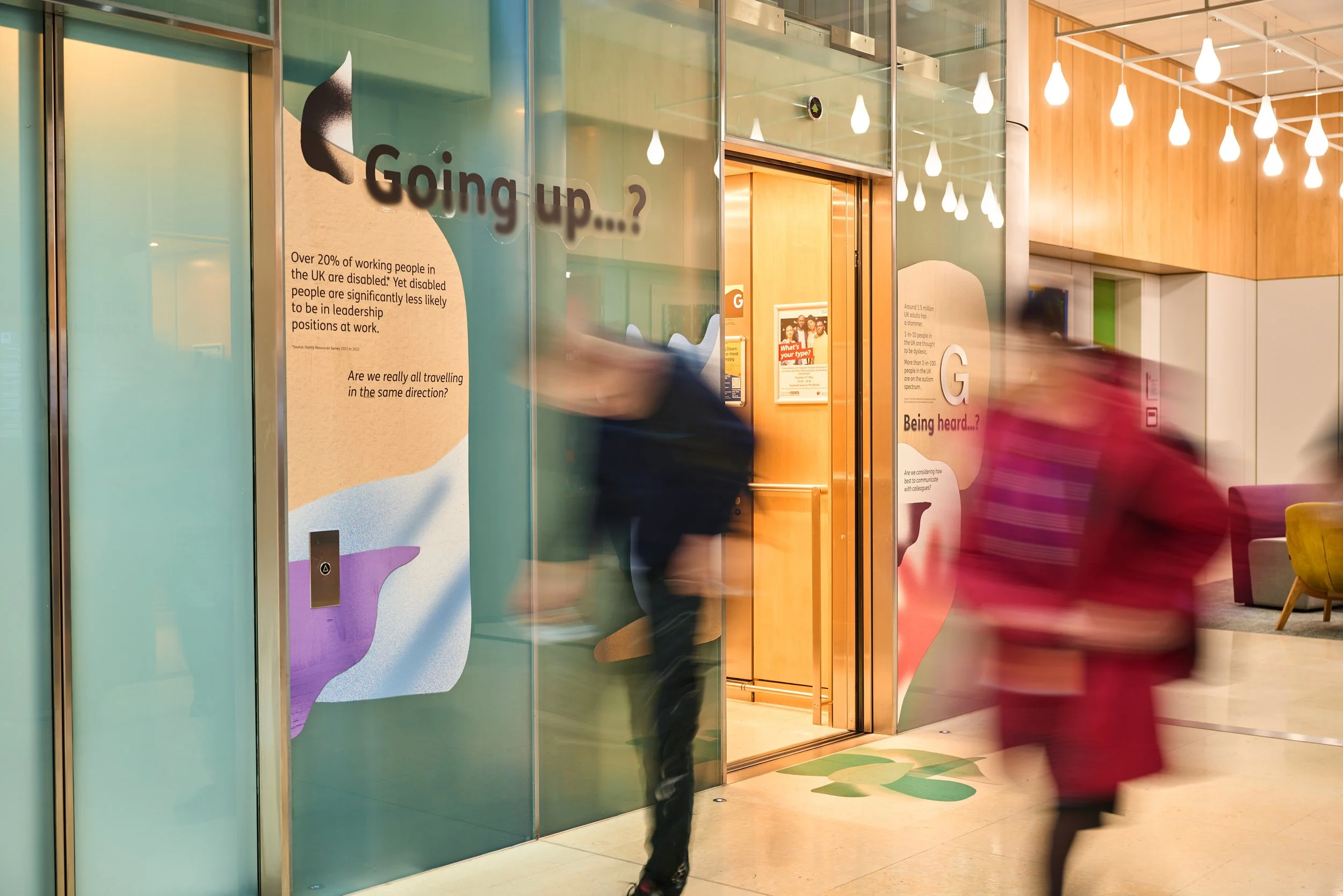 People entering an office building with informational posters on the glass walls, including questions like 'Going up...?' and 'Being heard...?'. The interior has warm lighting and modern decor.