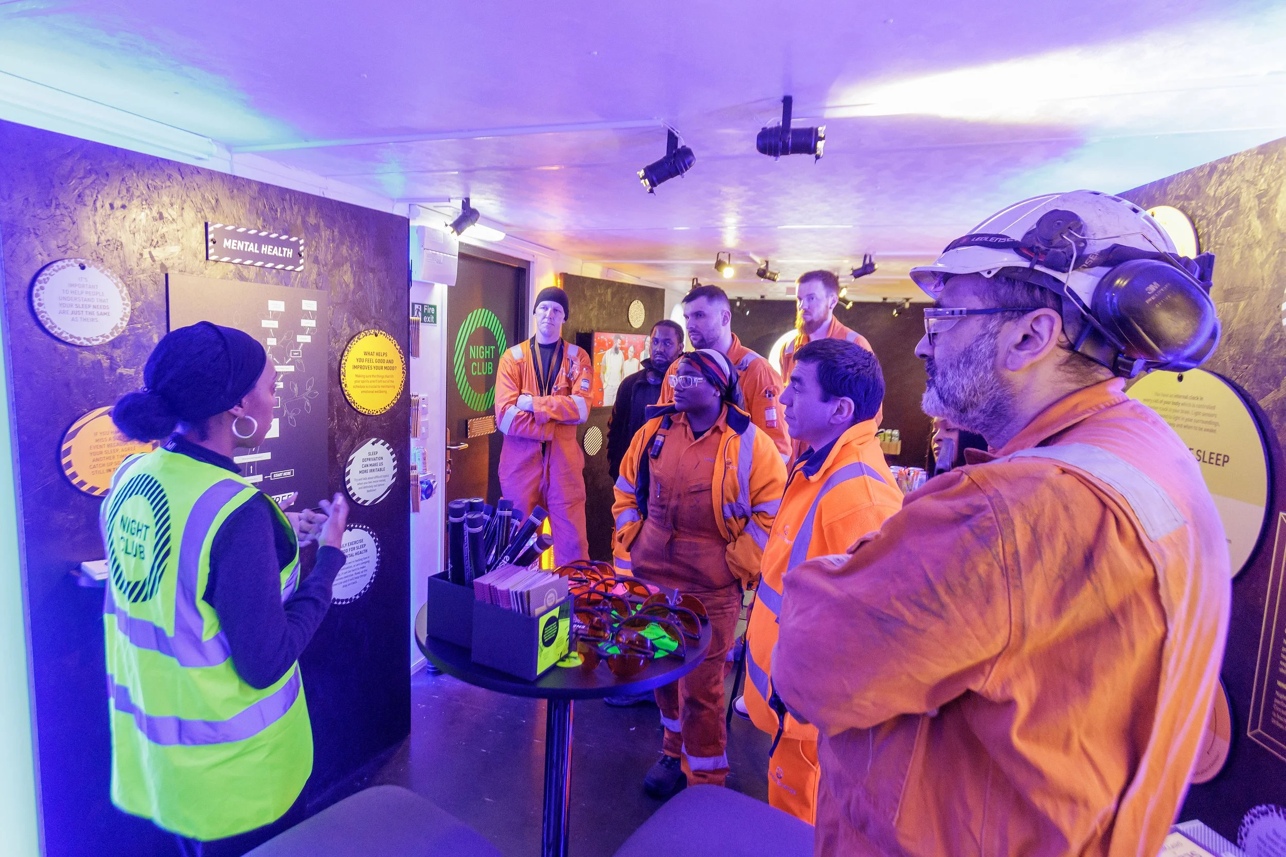 A woman is giving a presentation to a group of people dressed in orange uniforms in a room decorated with neon lights. The room has signs related to mental health and a table with safety glasses.