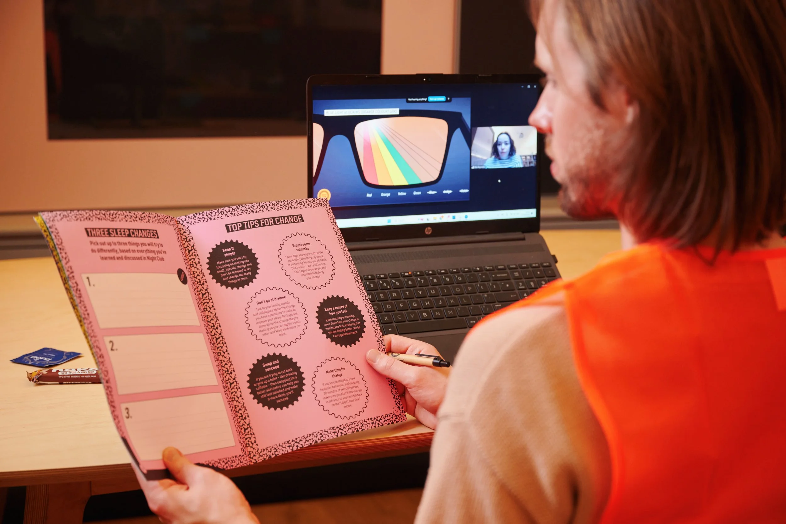 Person reading a pink booklet with tips for change at a desk in front of a laptop during a video call.