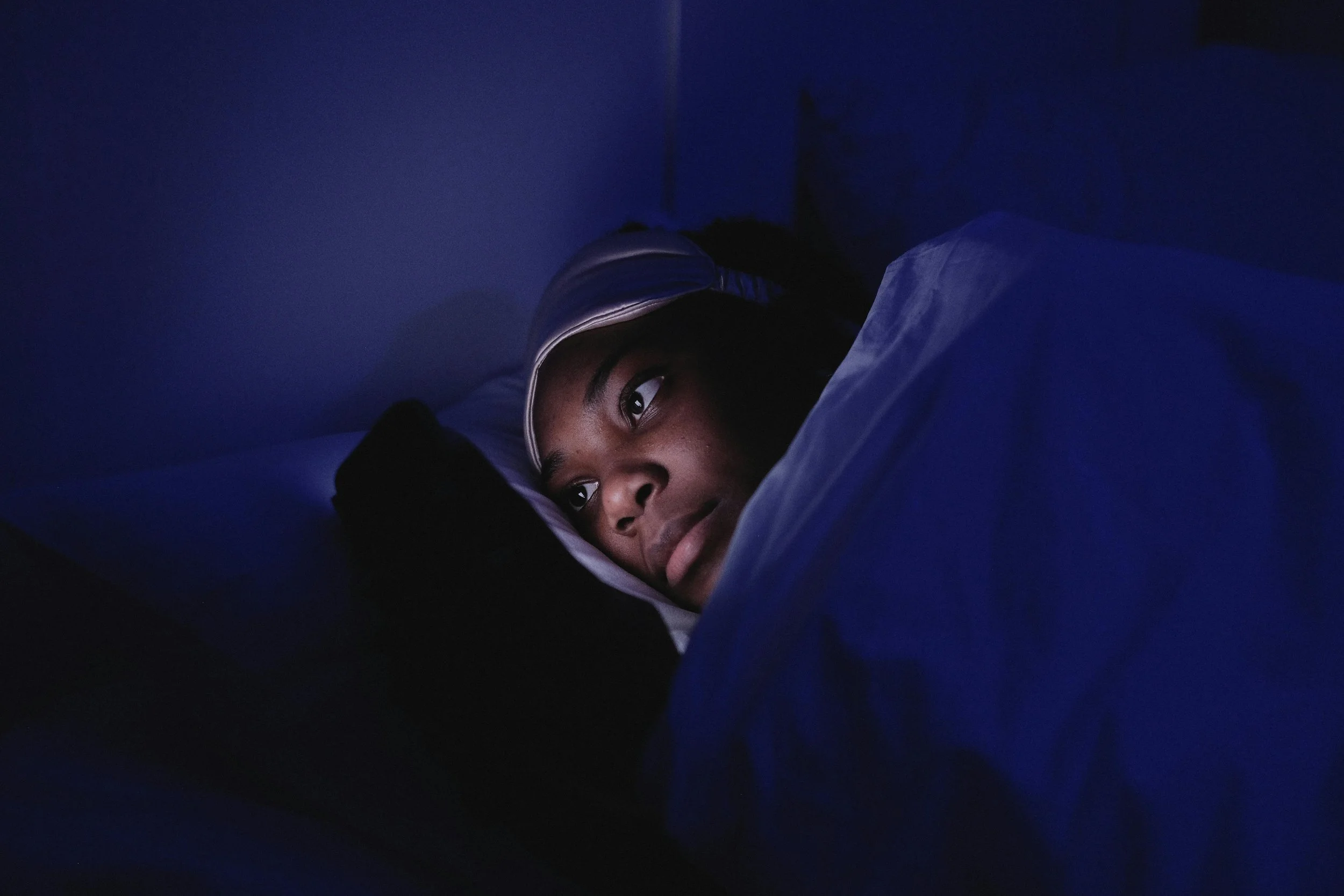 A person lies in bed awake looking at a phone screen with an eye mask on their head