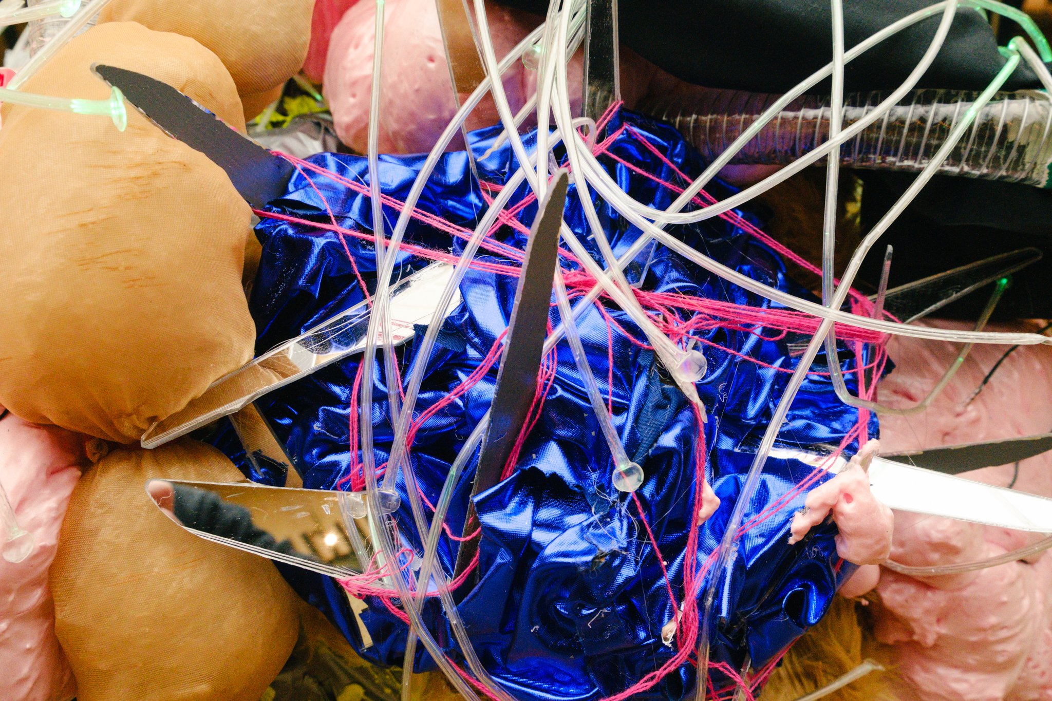 An abstract sculpture made of various materials, including bright blue fabric, pink thread, clear plastic tubes, and mannequin heads, all intertwined and layered in a chaotic arrangement.