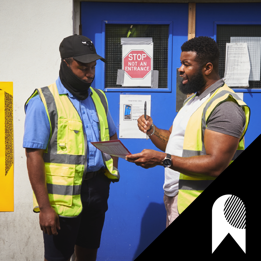Two men in yellow hi-vis vests standing outside a blue door, engaged in conversation. One man is holding a clipboard. 