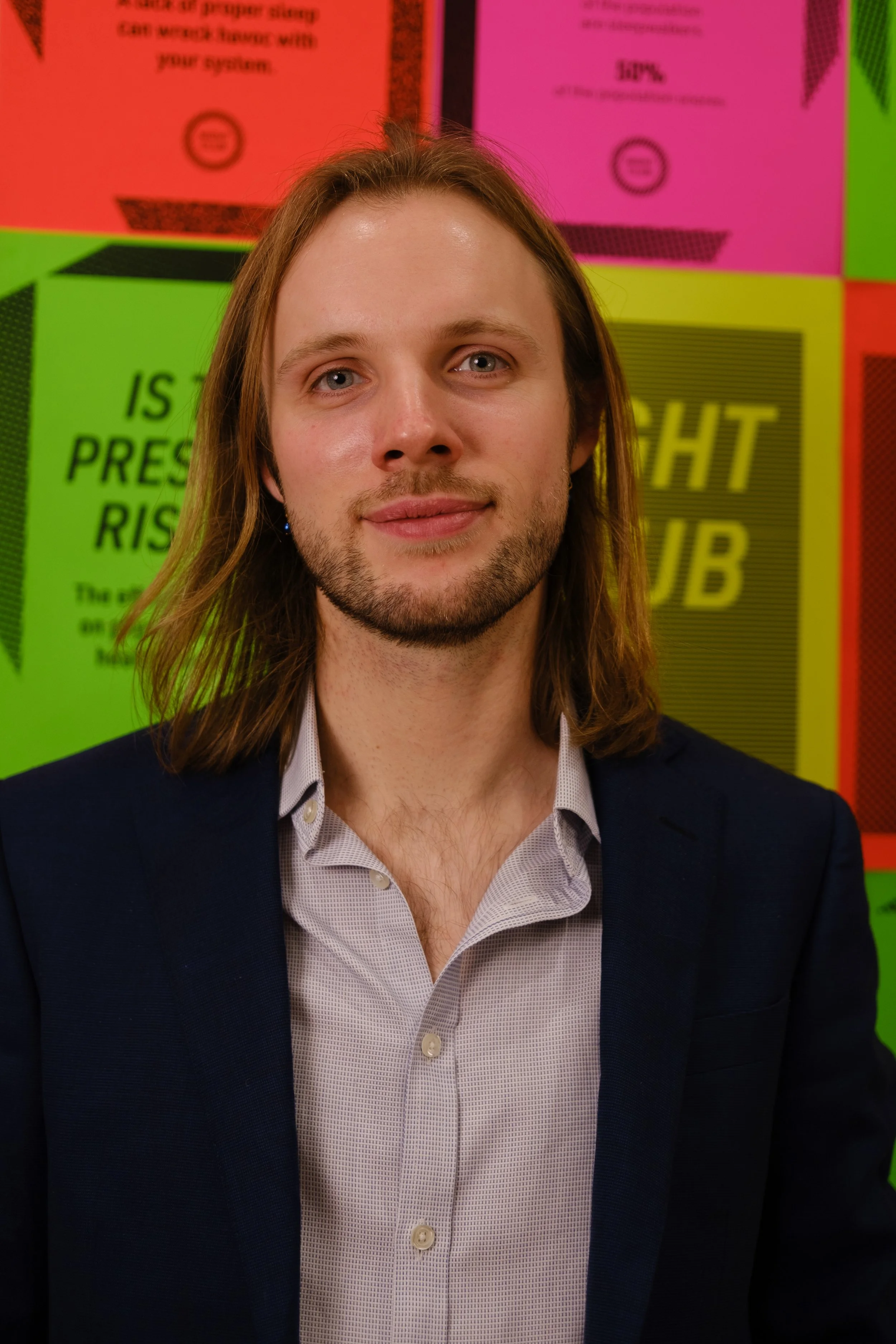 A photo of Finn Paul, Night Club Project Manager. A man with shoulder-length brown hair, a light beard, and light skin, wearing a white patterned shirt and dark blazer, standing in front of a colourful background with various posters.