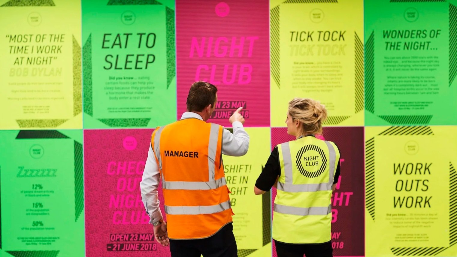 Two people, one man and woman, in safety vests labeled 'MANAGER' and 'NIGHT CLUB,' are looking at a colorful wall display advertising events, with the man pointing at one of the posters.