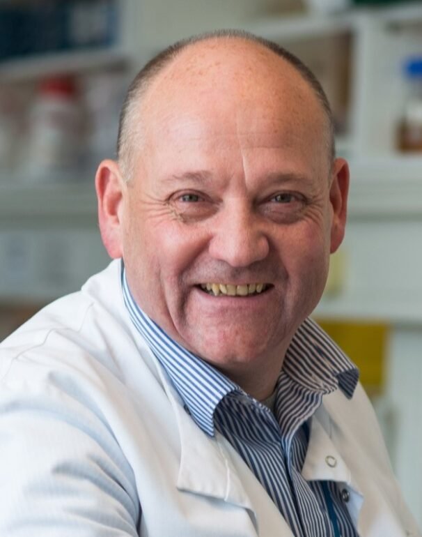A photo of Professor Russell Foster. A smiling man in a white lab coat and a blue striped shirt, in what appears to be a laboratory setting.