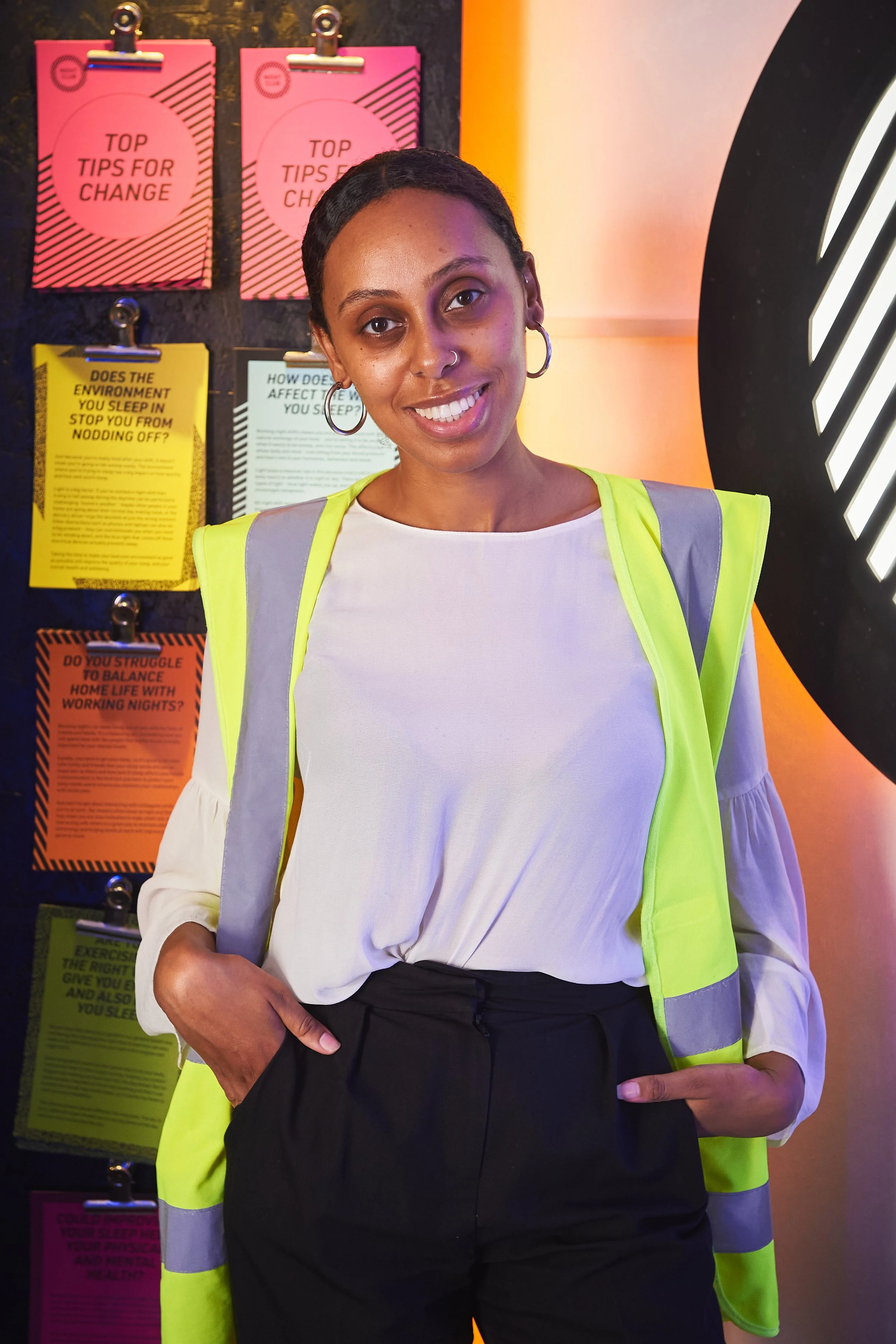 A photo of Ariam Abraham. A woman wearing a white shirt and a high-visibility safety vest standing in front of a colorful, informational poster display. She is smiling with her left hand in her pocket and has short dark hair and hoop earrings. 