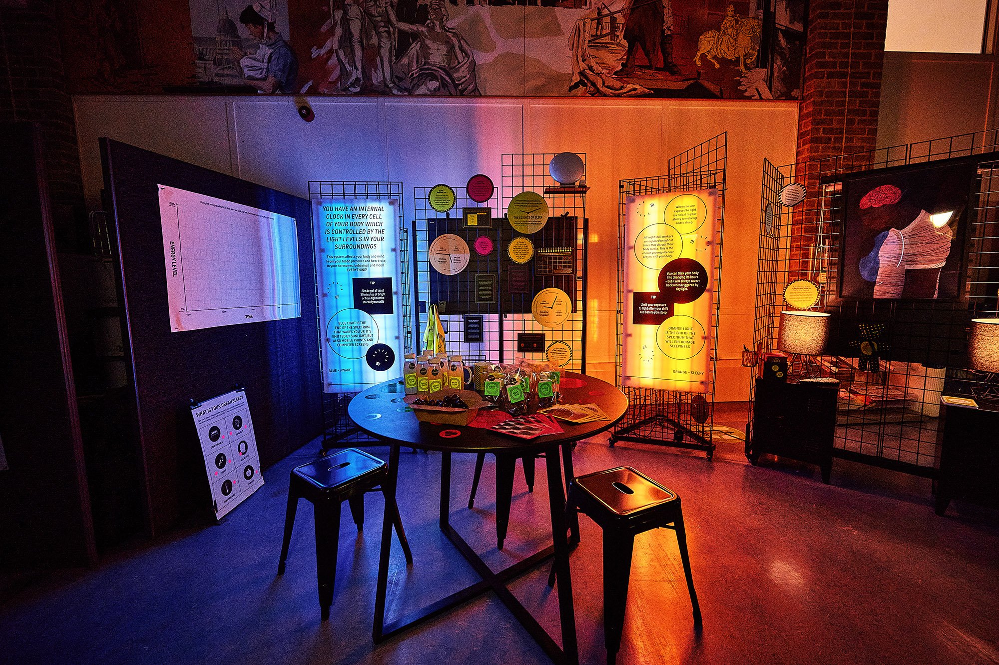 An indoor exhibit with illuminated informational panels, a round table with various items including bottled drinks and pamphlets, and black stools. The background features wire grid displays with colorful signs and artworks, and a brick wall is partially visible.