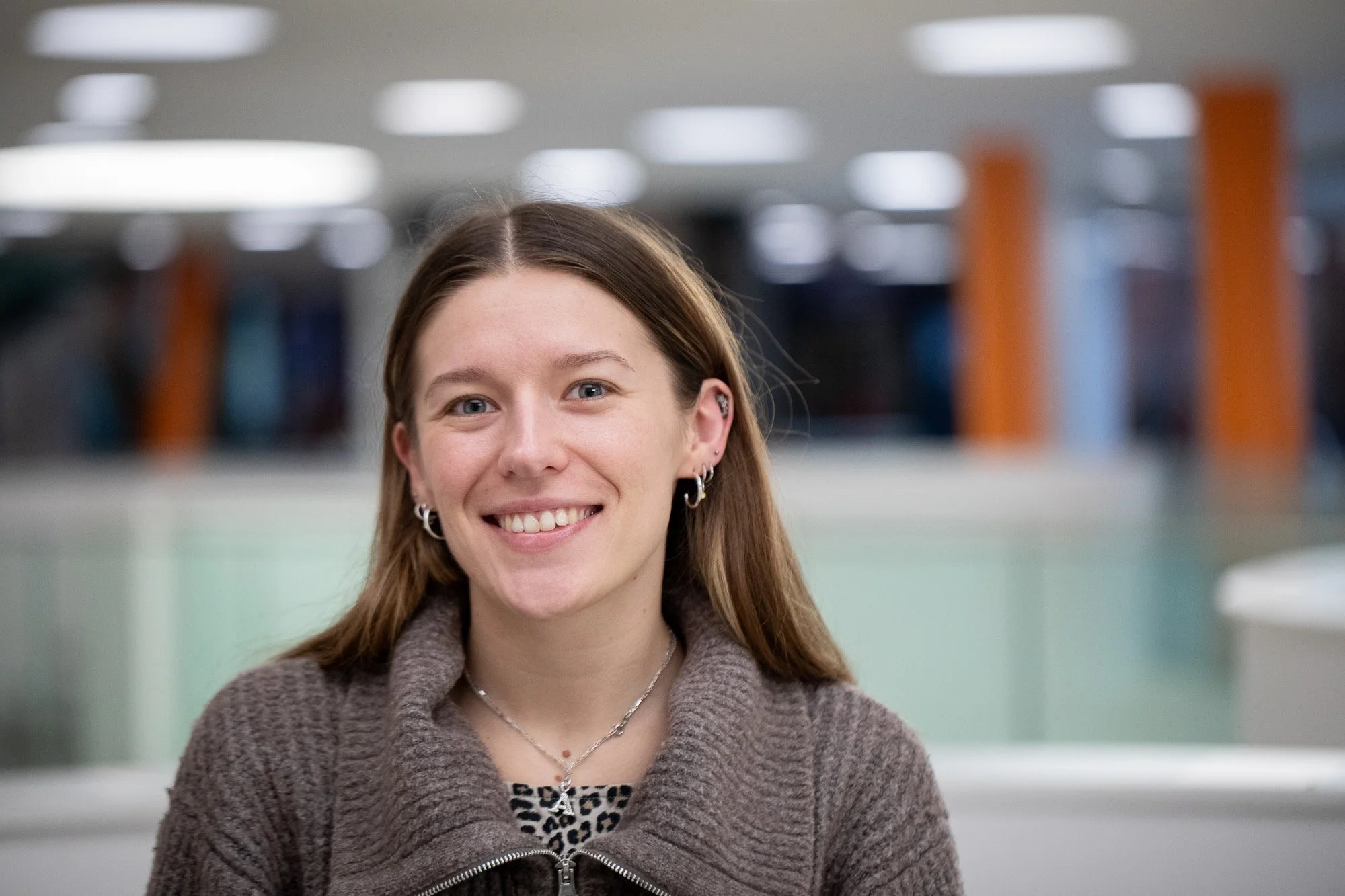 Smile of a young woman with long brown hair in an indoor setting with blurred lights and columns in the background.