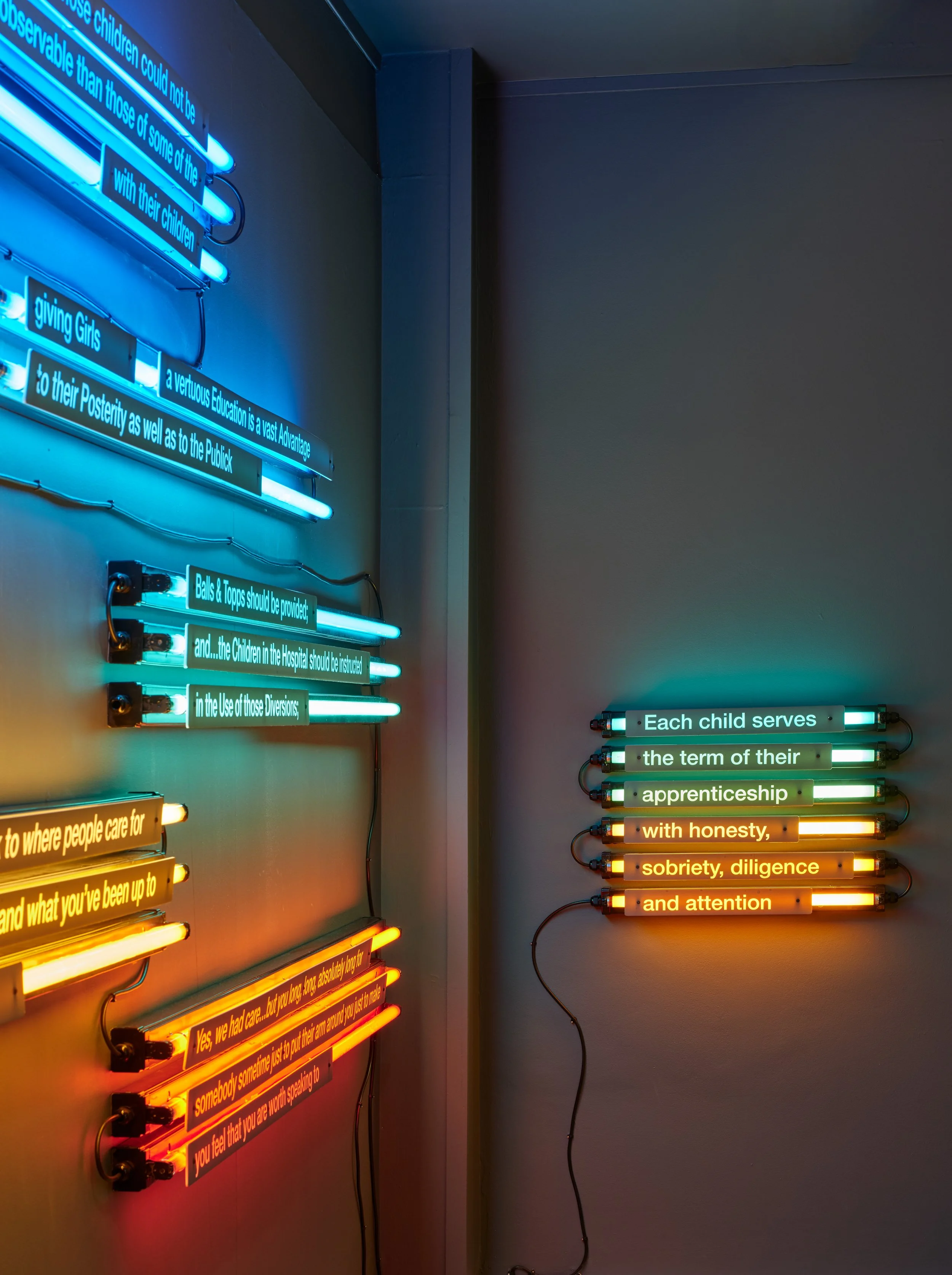 Neon wall art with various quotes in different colors, including blue, green, and orange, displayed on a gray wall.