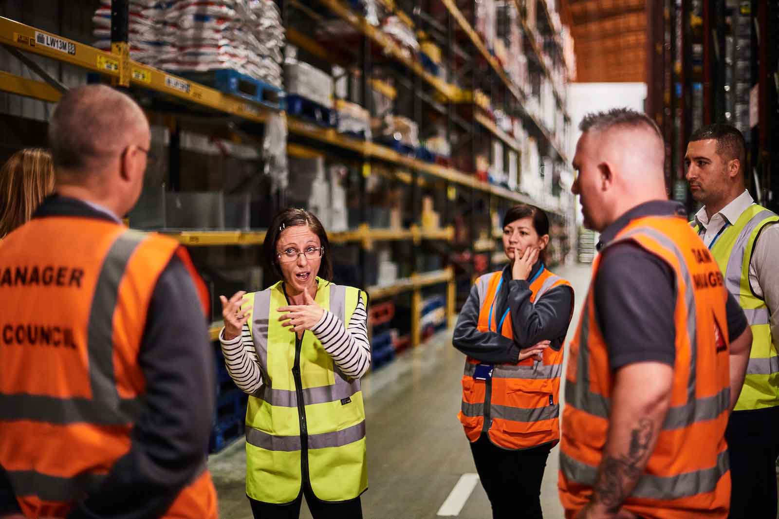 Group of six warehouse workers and managers having a discussion in a warehouse aisle with shelves of stored items. The woman in the center is gesturing while speaking.