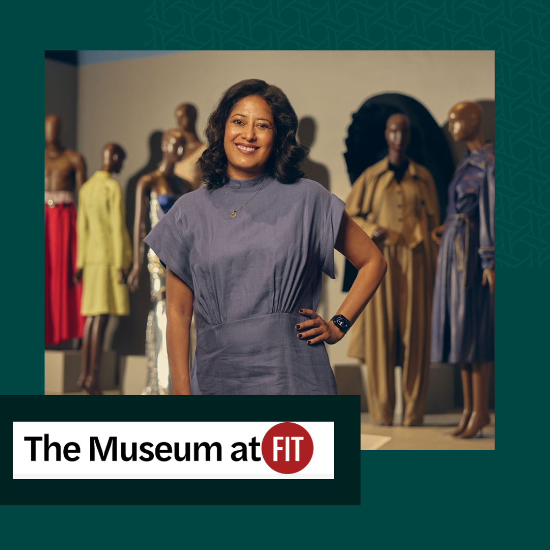 A woman stands with her hand on her hip, smiling at the camera, in a museum exhibit featuring mannequins dressed in colorful and metallic clothing.