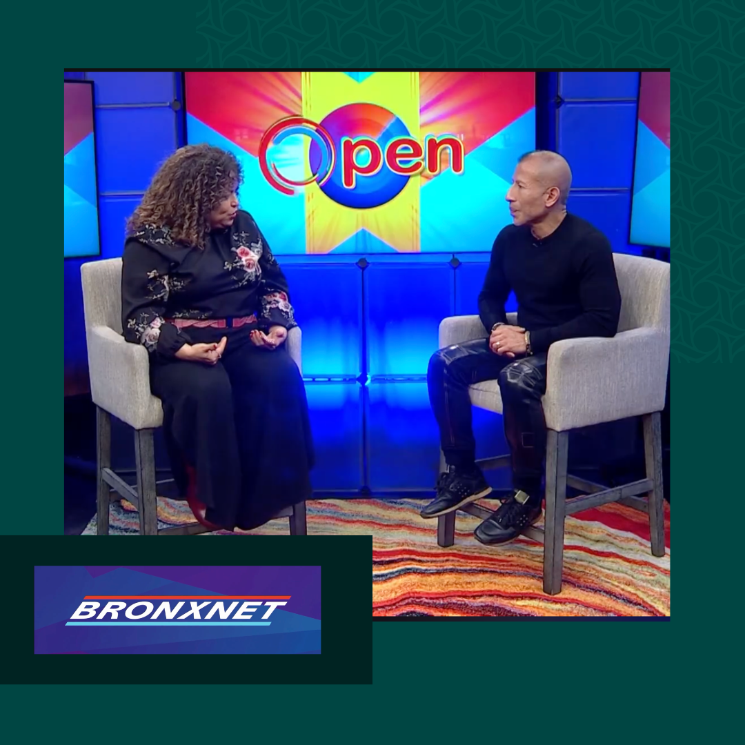 A television interview set with two people seated on chairs. Behind them is a colorful screen displaying the word 'Open' with a logo. A smaller logo in the corner says 'BronxNet.'