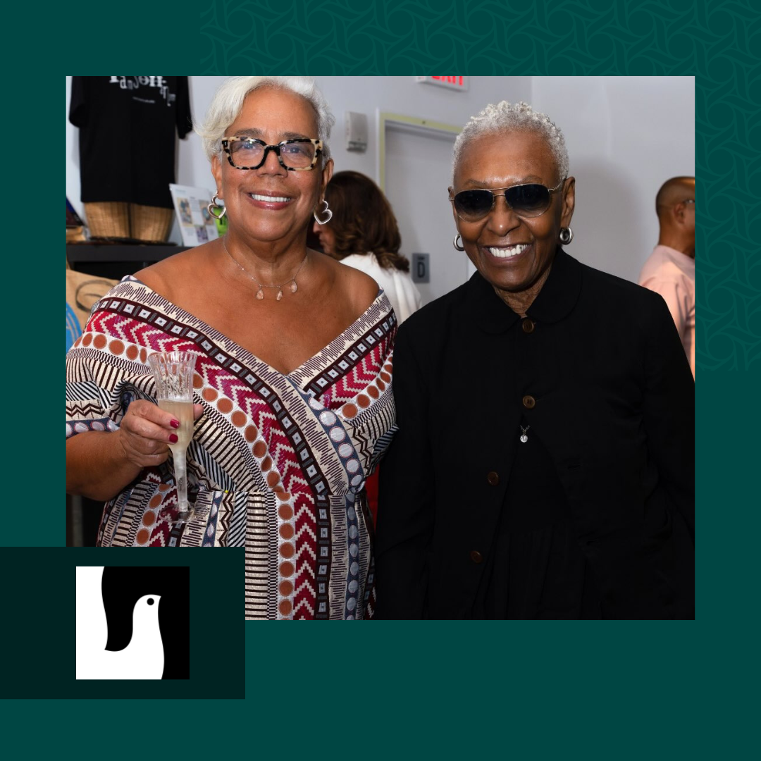Two women smiling and posing for a photo at a social event. One woman holds a glass of champagne, and the other is wearing sunglasses.