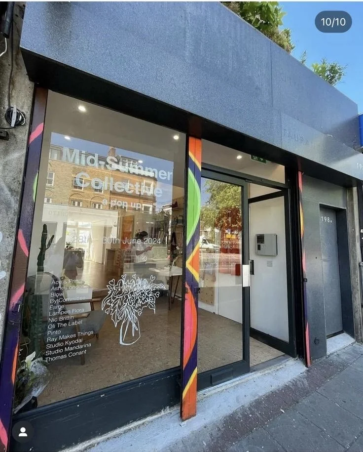 The storefront window with an advertisement for a mid-summer collective opening in June 2024, displaying a list of names and a white flower vase illustration inside.
