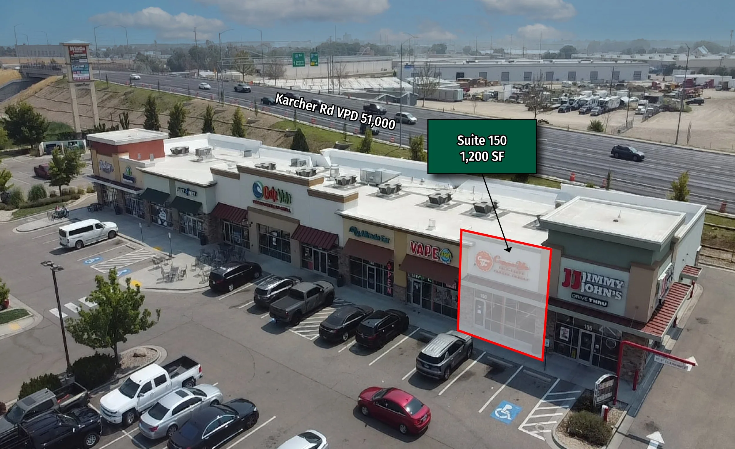 An aerial view of a shopping center parking lot with multiple cars, a row of small trees, and storefronts. A highway runs behind the shopping center, with several vehicles and green signboards. A green sign indicates a commercial space for lease, highlighting Suite 150 with 1,200 square feet.