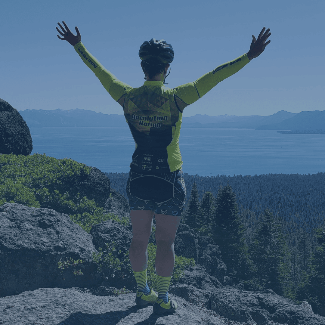 Marissa, a woman in cycling kit, stands at top of mountain with hands raised above head, overlooking a lake and evergreen trees
