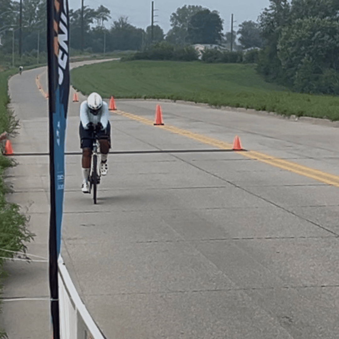 A woman, Lisa, with a white helmet and white cycling shirt and black cycling shorts bikes on a road toward a flag with cones and greenery in background