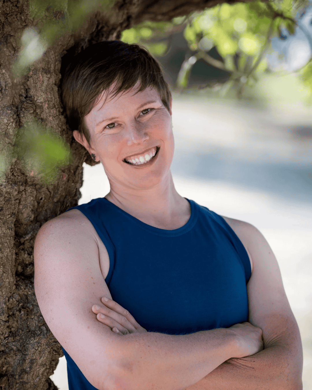 A photo of a woman, Marissa, with short brown hair is smiling and leaning back against a tree with a blue tank top