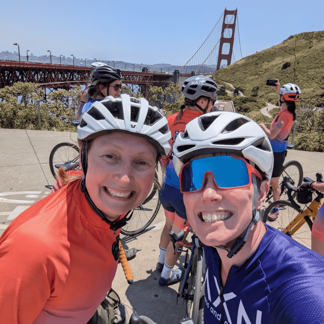 A woman in orange cycling shirt and white helmet, Alana, is smiling next to another woman, Marissa, in a white helmet and blue cycling shirt in front of the Golden Gate Bridge