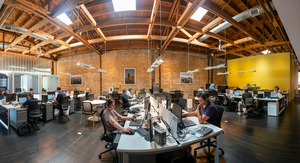 Open office with brick walls, rafters and skylights. QA Analysts test for accessibility and user experience.  Many sit at multiple desktop screens, some with headphones, and some with multiple devices to test connectivity.