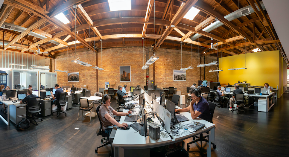 Open office with brick walls, rafters and skylights. QA Analysts test for accessibility and user experience. Many sit at multiple desktop screens, some with headphones, and some with multiple devices to test connectivity.