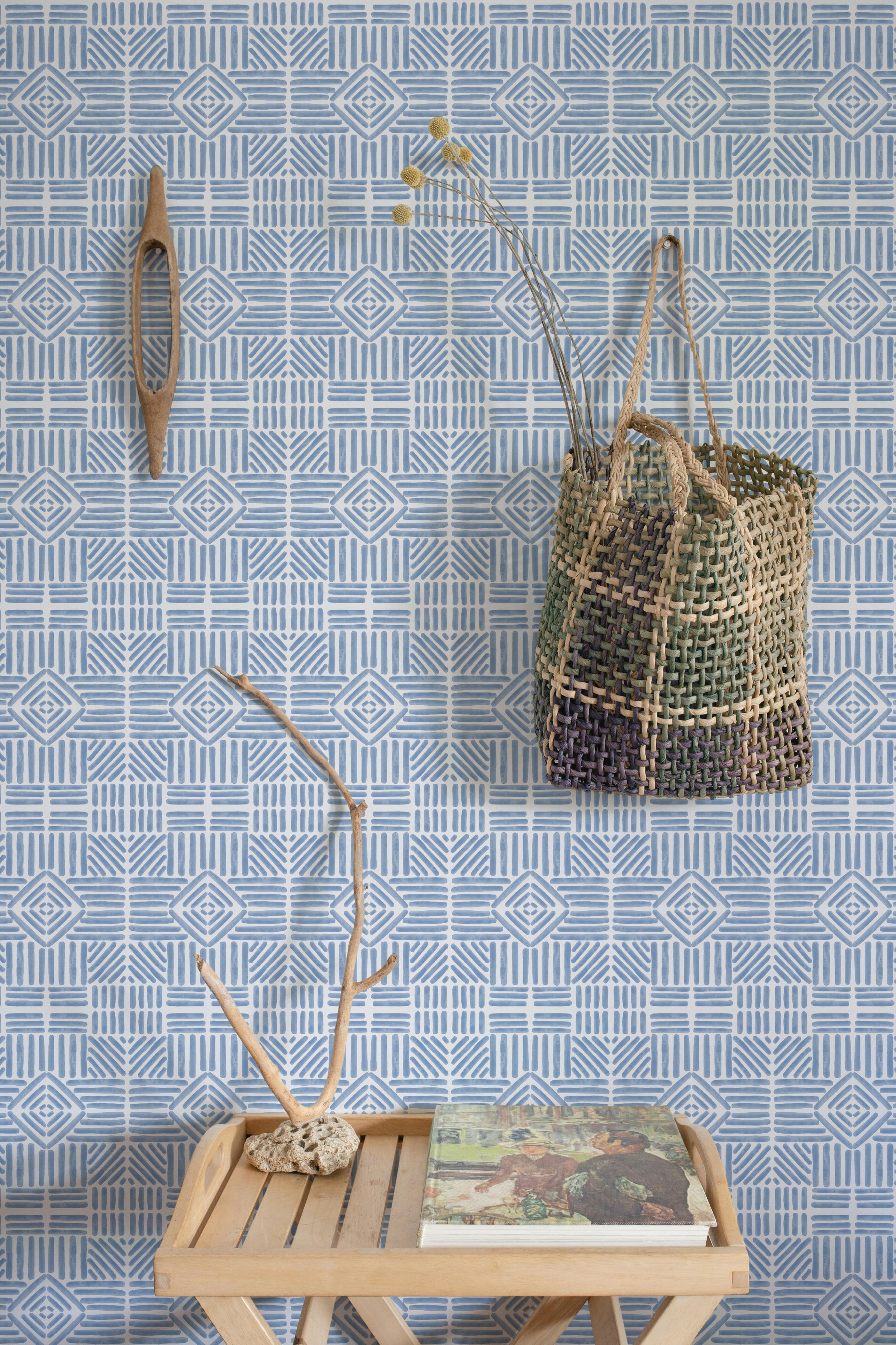 Coastal inspired blue geometric wallpaper with hand painted diamond and stripe motifs styled in a bright entryway. A woven market tote, driftwood accents, and natural wood furniture highlight the fresh, relaxed feel of the pattern.