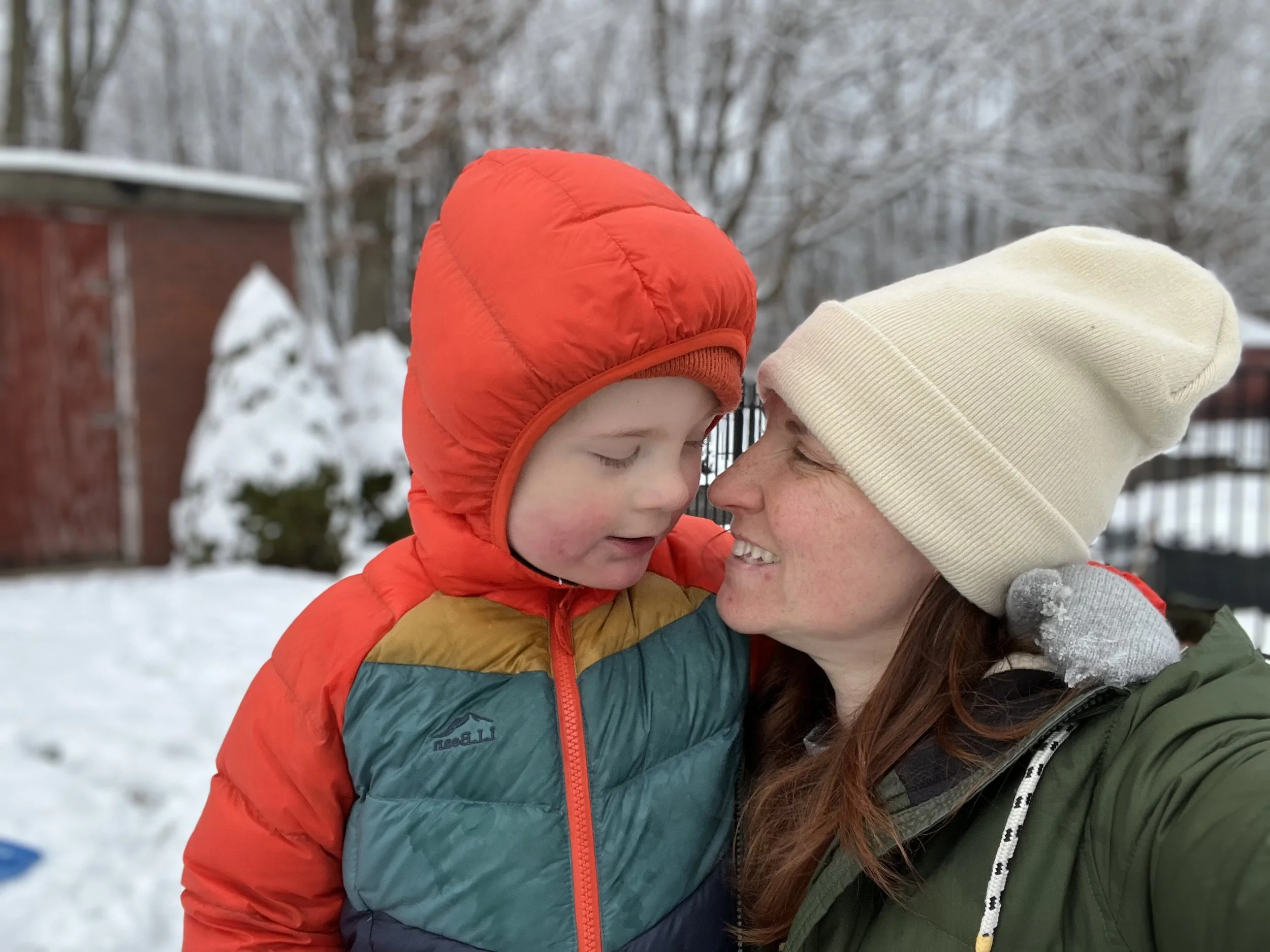 A mom and her son bundled up in winter coats and hats sharing a hug looking at each other outside in the snow.